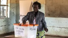 One woman in a patterned jacket and African wrap for a skirt stands at a ballot box  putting her vote in. She is in an empty classroom and a blackboard can be seen behind her.