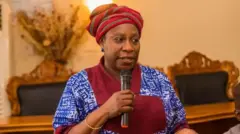 Sẹnetọ Iyabo Obasanjo-Bello. O we gele alawọ pupa, wọ aṣo alawọ aro to ni awọ pupa ninu, o si mu ẹrọ gbohungbohun dani nigba to n sọrọ. 