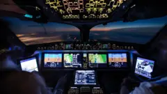 A imagem mostra o interior de uma cabine de comando moderna durante um voo ao amanhecer ou entardecer. Os painéis de instrumentos e telas digitais iluminam o cockpit com tons de azul, verde e amarelo, exibindo informações de navegação, altitude e sistemas da aeronave. Em primeiro plano, é possível ver parcialmente dois tripulantes sentados diante dos controles. Pela ampla janela frontal, observa-se o horizonte com o céu em tons azulados e alaranjados acima de uma camada de nuvens, criando uma atmosfera tecnológica e ao mesmo tempo contemplativa.