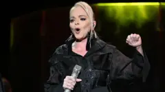 Женщина со светлыми крашеными волосами и в макияже стоит на сцене в черном костюме и микрофоном в руке. Судя по мимике, женщина что-то кричит зрителям, поднял кулак левой руки.