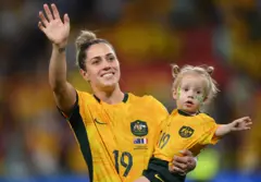 Katrina Gorry carries two-year-old daughter Harper, while wearing matching football kits, as they wave to the crowd
