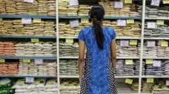 A woman wearing a blue printed top stands in front of a grocery store shelf stocked with pulses and grains