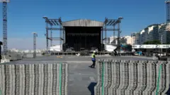 Uma grande estrutura de palco em montagem à beira-mar, cercada por prédios residenciais ao fundo e um céu limpo e azul; no primeiro plano, há pilhas organizadas de blocos de piso, enquanto um trabalhador com capacete e colete refletivo caminha pelo espaço amplo ainda em preparação, ao lado de equipamentos, andaimes e torres metálicas.