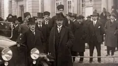 İsmet İnönü ve diğer delegeler bir otomobil önünde kameraya poz veriyor. Üzerlerinde takım elbise, palto ve uzun şapkalar var.