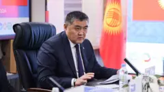 Ташиев в синем костюме сидит за столом на фоне флага Кыргызстана