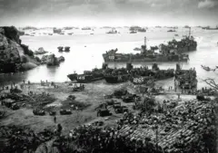 Vista desde lo alto de embarcaciones militares estadounidenses fondeadas en la costa de la isla de Okinawa. En tierra, hay varios vehículos, hombres y bultos amontonados.