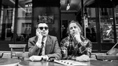 Dois homens com o braço apoiado sobre uma mesa ao ar livre em foto em preto e branco