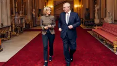 Emily Maitlis berjalan bersama Andrew melewati Istana Buckingham. Mereka berada di atas karpet merah dengan dinding marmer dan patung-patung. Sebuah kursi panjang berwarna merah membentang di sisi koridor.