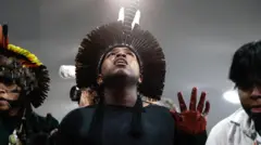 抗議に参加する先住民の人たち。中央の男性は上を仰いでいる。羽で作った頭飾りをして、手には赤い染料をつけている