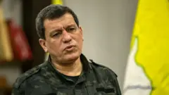 Abdi kamuflaj kıyafetleriyle konuşuyor, arka planda SDG bayrağı kısmen görünüyor. 