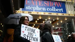 A imagem mostra um grupo de pessoas em frente ao teatro do programa “The Late Show with Stephen Colbert”, à noite e sob chuva. Em primeiro plano, uma mulher segura um guarda-chuva e um cartaz com a frase “Thank You Stephen”, ou “obrigado, Stephen”, em português. Ao fundo, a marquise iluminada do programa aparece em destaque, cercada por luzes amarelas e pela movimentação da rua.