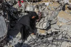 سيدة فلسطينية محجبة ترتدي لباساً أسود تمسك بيدها مطرقة وتجلس على أنقاض منزلها. يظهر وجهها من الجانب. الصورة التقطت نهاراً.