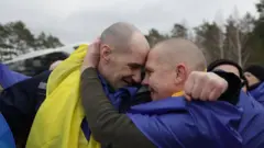 Двоє людей, загорнутих у синьо‑жовті прапори, обіймаються просто неба. Один із них тримає іншого за голову, притягуючи ближче. Навколо видно інших людей у подібному вбранні, а тлом слугує похмуре зимове небо та ліс.