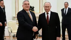 Dwóch mężczyzn w ciemnych garniturach ściska sobie dłonie w formalnym, ozdobnym pokoju, podczas gdy dwóch innych stoi za nimi przy białych krzesłach i ozdobnym kominku.