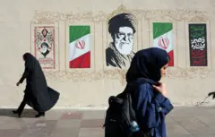 Dos mujeres con velo caminan por la acera frente a un mural que tiene banderas iraníes, caligrafía decorativa y un gran retrato de Alí Jamenei. Una mujer lleva una túnica negra, mientras que la otra viste un abrigo azul oscuro y lleva una mochila.