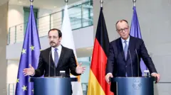 Kıbrıs Cumhurbaşkanı Nikos Hristodulidis ve Almanya Başbakanı Friedrich Merz Berlin'de basın toplantısında kürsüden konuşuyor. İki lider koyu renk takım elbise giymiş ve arkalarında AB ve Almanya bayrakları var