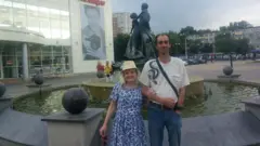 Sergei e Tatyana estão em uma praça, em frente a uma fonte. Eles olham para a câmara. Ela é loira e usa um vestido de flores azul e um chapéu. Ele está com uma camisa branca e calça jeans.