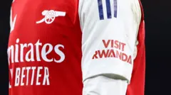Umwenda wa Arsenal w'umutuku n'umweru wanditseho amagambo Visit Rwanda ku kaboko