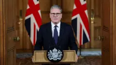 صورة أرشيفية لرئيس الوزراء البريطاني، كير ستارمر وهو يقف على منصة خطاب داخل 10 داونينج ستريت، وبجانبه علمي المملكة المتحدة. - صورة من الأرشيف