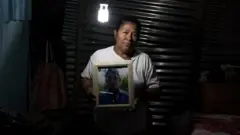 Una mujer sostiene el retrato de su hijo adulto. Está en un cuarto de una casa humilde, a media luz.