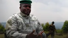 Umugabo wambaye imyenda yijimye ya gisirikare n'ingofero y'icyatsi kibisi, akoze ikimenyetso n'ibiganza, inyuma ye hari umugabo wambaye imyenda yijimye, n'imisozi inyuma kure. 
