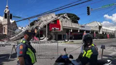 Policiais uniformizados estão em frente a uma estrada barricada. Atrás deles está um prédio parcialmente destruído com uma placa vermelha do Jollibee.