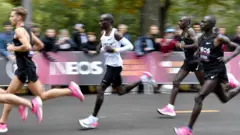 A imagem mostra o queniano Eliud Kipchoge com uma camisa branca correndo durante um evento especial de maratona realizado em Viena em outubro de 2019. Ele está cercado por corredores de elite vestindo preto, que ditam o ritmo e ajudam a criar um efeito de arrasto similar ao que ocorre em uma corrida de Fórmula 1.