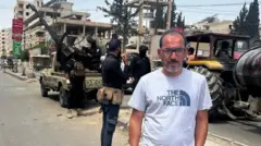 Ферас Килани, седеющий мужчина в очках, стоит на фоне техники сирийских сил безопасности