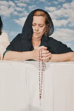Una mujer con un chal negro reza al aire libre, sosteniendo un rosario.