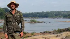 Rodrigo em pé, sorrindo, em frente a aparentemente uma lagoa ou rio