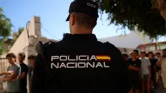 Um policial aparece em primeiro plano, de costas para a câmera, enquanto crianças fazem fila do lado de fora de uma delegacia para serem registradas pelas autoridades.