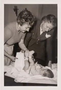 Cristina segura os pés do bebê com uma das mãos, enquanto com a outra segura uma fralda. María Luisa a ajuda a segurá-los. As duas olham para o bebê. Cristina veste uma camisa de mangas curtas e uma saia, e María Luisa usa um tailleur escuro