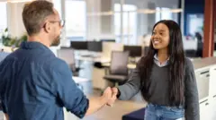 A young woman wearing a grey jumper over a shirt and blue jeans shaking hands with a man wearing a blue shirt and glasses in an office 