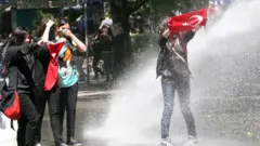 Polisin su püskürtüğü kadın protestoculardan biri elinde Türk bayrağıyla yüzünü kapatıyor, diğer üçü bir arada duruyor.