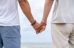 Um casal homossexual de mãos dadas em frente à praia.