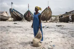 Mace a gefen teku, dauke da buhu a hannunta a gaban wani kwale-kwale, sanye ta ke da  bular riga da adiko ruwan dorawa.