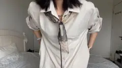 Uma mulher com cabelo na altura dos ombros, vestindo uma camisa larga de cor esbranquiçada, um cachecol preto e creme com estampas ao redor do pescoço, está em pé diante de uma cama.