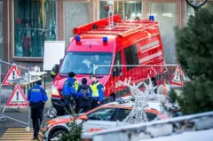 Un grupo de policías conversan frente a una ambulancia roja y un automóvil de la policía en una calle que ha sido acordonada. Se ven unos pinos y adornos navideños