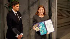 Ana Corina Sosa Machado, hija de María Corina, sostiene en sus manos la medalla del nobel de la paz y un certificado. sonríe. viste ropa gris, tiene cabello hasta los hombros. A su lado está Jorgen Watne Frydnes, quien sonríe. Viste un traje y corbata.