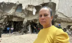 Laura Barrios está de pie frente a un edificio mientras busca los restos de su cuñado. Lleva un suéter amarillo y su cabello oscuro está recogido.