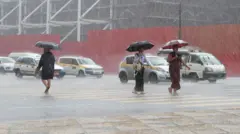 Tres personas cruzan una carretera bajo una lluvia intensa en Myanmar, con tráfico al fondo. Todas llevan paraguas mientras la lluvia rebota en el paso de peatones.