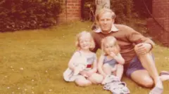 Foto antiga de família mostra Smyth vestindo bermuda azul e suéter marrom, sentado na grama ao lado das duas filhas pequenas. Parece ser um dia quente, e todos sorriem para a câmera