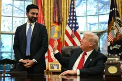 Mamdani is seen standing next to Trump, who is seated behind his desk in the Oval Office. Both are smiling and Trump is reaching out to jokingly tap him on the arm