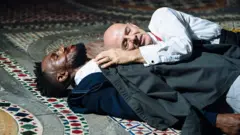 Casal de atores no palco: um homem negro de barba e terno está deitado no chão, com sangue no rosto, enquanto um homem branco e calvo de camisa se deita sobre seu peito