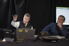 Un funcionario hace un gesto con la mano mientras mira hacia delante, y Hegseth mira atento con su mano izquierda sobre la parte inferior de su cara.