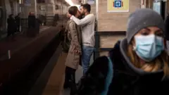 A couple kissing on an underground platform in Italy as a woman wasks past wearing a mask