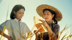 画面中两名女性站在田间，其中一人正向另一人念出一封信的内容。