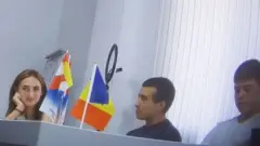 Узун жигарранг сочли аёл (Алина Жук) атрофини ўраб олган гуруҳга жилмайиб қарайди — улар катта стол атрофида ўтиришган, фонда иккита байроқ кўриниб турибди, улардан бири қизил, сариқ ва кўк рангли Молдова байроғи.