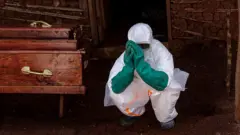 Un agent de santé en équipement de protection individuelle complet - costume blanc et gants en caoutchouc verts - accroupi à droite d'un cercueil en bois aux poignées dorées