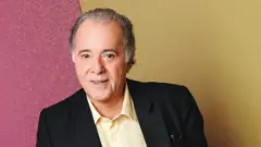 Tony Ramos, com cabelos grisalhos penteados para trás, aparece em um retrato posado. Ele veste blazer preto sobre uma camisa clara e olha diretamente para a câmera com expressão simpática. O fundo é dividido em duas cores — rosa à esquerda e amarelo à direita.